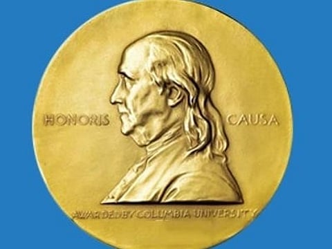 Gold Pulitzer Prize medallion featuring a side profile of Benjamin Franklin, with "Honoris Causa" text. Below, "The Pulitzer Prizes" is written in white on blue.