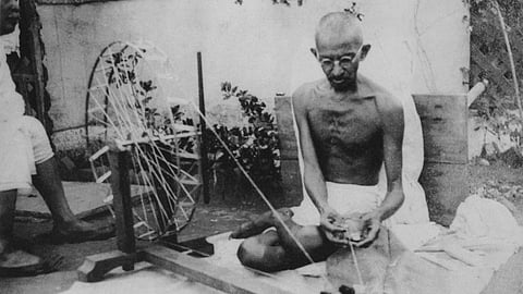 Mahatma Gandhi seated cross-legged on the ground, spinning cotton using a charkha. He appears focused, conveying simplicity and self-reliance in a serene setting.
