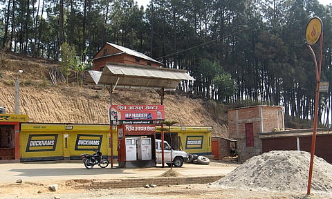 Rural gas station with a motorcycle and car beside a fuel pump. Yellow storefronts and wooded hillside in the background, creating a tranquil scene.