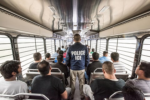 backview of people sitting in a bus while a man waring a 'Police ICE' jackets stands in the middle
