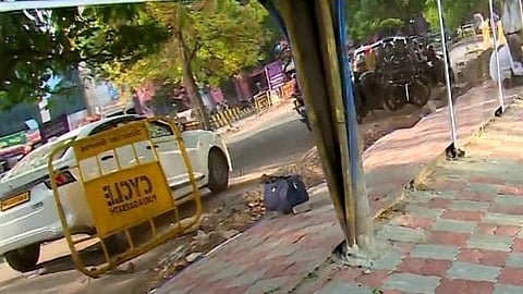 A reflective glass fence alongside a tree-lined street shows a distorted view of traffic and surroundings, including a car, barricade, and a bag.