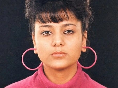 A woman with a high bun and bangs wears large pink hoop earrings and a matching turtleneck. She gazes forward with a neutral expression against a dark backdrop.