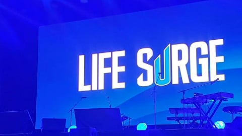 Blue-lit stage with "LIFE SURGE" in bold white letters on a large screen. Silhouettes of musical equipment, creating an energetic atmosphere.