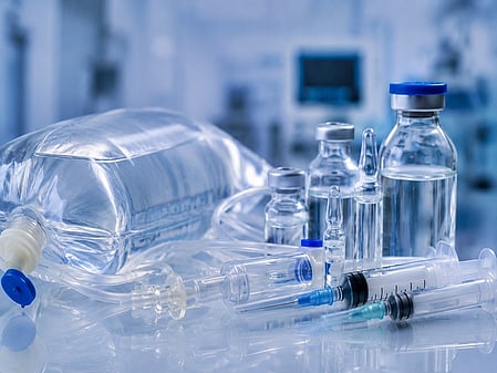 A variety of medical supplies on a table, including syringes, vials, and an IV bag. The setting is a sterile, modern medical environment.