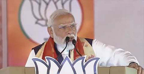 Image of PM Modi with grey hair and glasses speaks passionately at a podium adorned with a lotus symbol, set against an orange background with a blurred emblem.