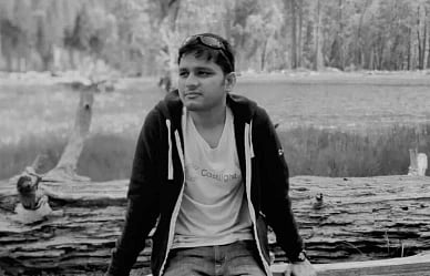 A person sits on a log in a scenic outdoor setting by a lake, wearing a hoodie and t-shirt. The background features trees, conveying a calm, reflective mood.