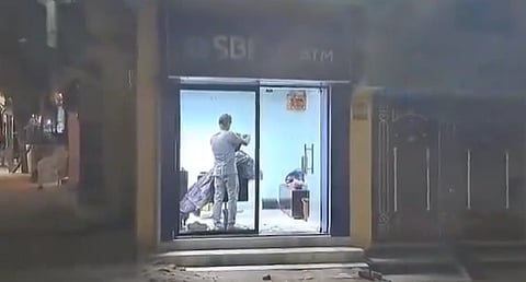 A group of people in an ATM booth at night, with one person crouched near the floor and another holding a bag, creating a tense atmosphere.
