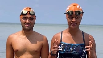 Two swimmers in orange caps and goggles stand by the ocean. One is shirtless, and the other wears a swimsuit, gesturing with hands. The mood is focused and determined.
