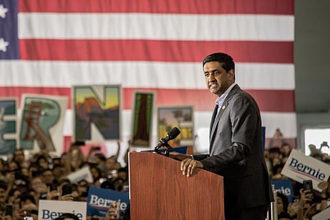 Ro Khanna at a podium with a microphone, set against a large American flag. Audience holds "Bernie" signs, creating an energetic atmosphere.