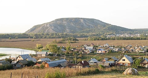 Rustic village with scattered houses, tin roofs, and a nearby pond. A large hill dominates the background, conveying tranquility and simplicity.