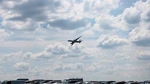 Aeroplane is shown flying in a cloudy sky below are cars parked