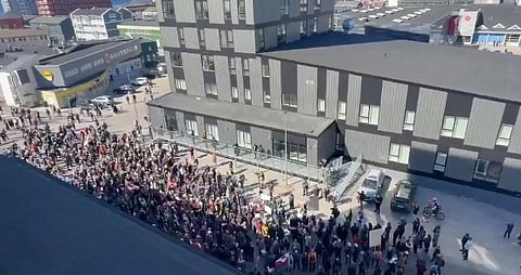 Crowd of people holding flags gather for a protest outside a large, gray, modern building. The scene is lively and energetic, under clear skies.