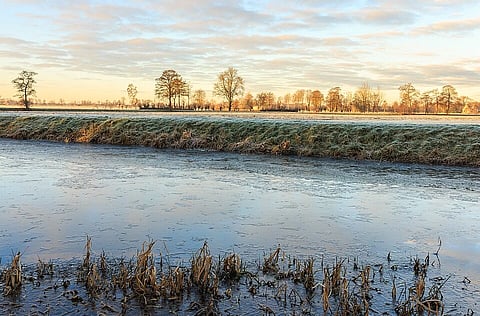 Frosty morning landscape with a frozen pond in the foreground, grassy bank, and leafless trees under a partly cloudy sky, conveying tranquil calmness.