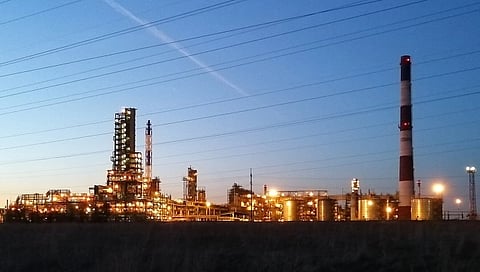 Industrial refinery at dusk with bright lights illuminating tall structures and a striped smokestack against a deep blue sky, conveying a serene yet industrious atmosphere.