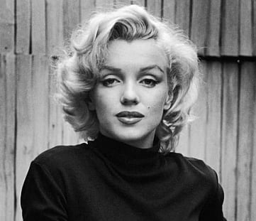 Black and white portrait of a Marilyn Monroe with short, wavy blonde hair and a black turtleneck. She gazes calmly at the camera, standing against a wooden fence background.