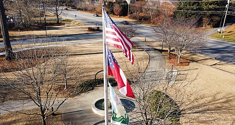 Aerial view of three flags, including the U.S. flag, fluttering above a circular plaza with leafless trees, intersecting paths, and a surrounding road.