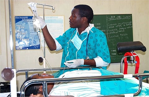A healthcare worker in teal scrubs adjusts an IV drip beside a patient on a hospital bed. The setting is a medical room with charts on the wall.