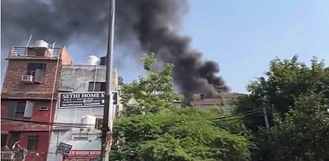 Thick black smoke rises from a building behind trees on a sunny day. Nearby, a sign reads "Sethi Home," creating a sense of urgency and alarm.