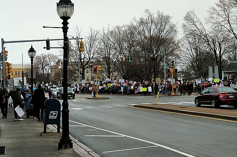 A large crowd gathers at a street corner, holding colorful signs in a peaceful protest. Overcast skies and bare trees set a somber tone. Crosswalks and lampposts frame the scene