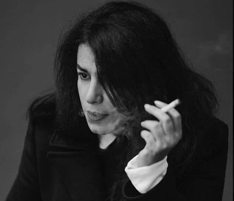 A black-and-white image of Marjane Satrapi with long, dark hair, gazing thoughtfully while holding a cigarette. The background is plain, conveying a contemplative mood.