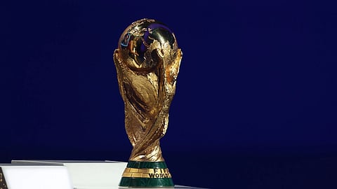 The image shows the FIFA World Cup trophy, a gold statue depicting two figures holding a globe, set against a dark blue background.