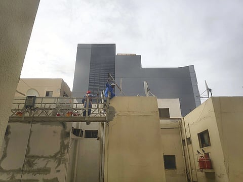 Two workers in safety gear stand on a suspended platform, painting a beige building. A modern gray building looms in the cloudy background.