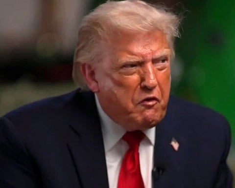 Image of Donald trump sitting in an interview and making a uneasy expression on his. he is wearing a blue suit with a badge of American flag