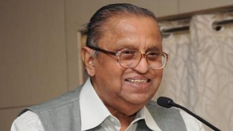Professor Rajaraman, a man in a grey vest and glasses, is smiling warmly at the camera.