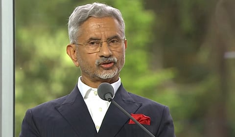 In the image EAM S Jaishankar is shown speaking in an international forum behind him green plants are shown outside a glass wall
