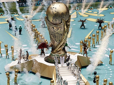 A large, gold World Cup trophy is surrounded by performers in elaborate costumes on a field. Water jets spray upwards, creating a festive and energetic scene.