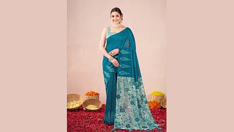 A woman in a teal saree stands on a red flower-petal carpet, smiling. Woven baskets with colourful flowers are in the background, evoking a festive atmosphere.