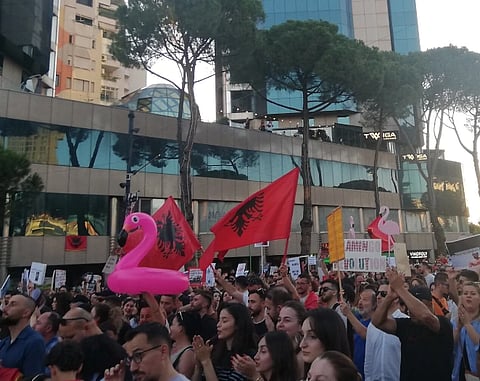 A large crowd gathers in front of modern buildings, holding red flags with black eagles and pink flamingo floats, conveying a lively protest atmosphere.