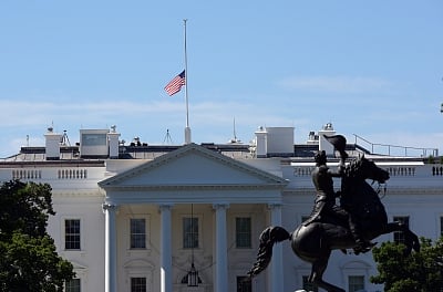 The image shows the White House with an American flag at half-mast. A silhouetted equestrian statue is in the foreground under a clear blue sky.