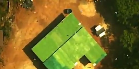 Aerial view of a green-roofed building on reddish-brown soil, surrounded by scattered trees and vegetation. The scene is calm and remote.