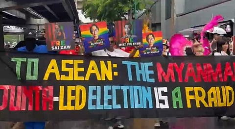 Protesters hold a banner reading "To ASEAN: The Myanmar junta-led election is a fraud" in bold colors. They also display signs with faces and call for freedom.