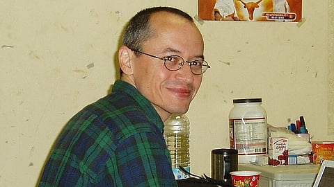 In the image comic journalist Joe Sacco is shown behind him are many boxes in a table and a poster in the wall