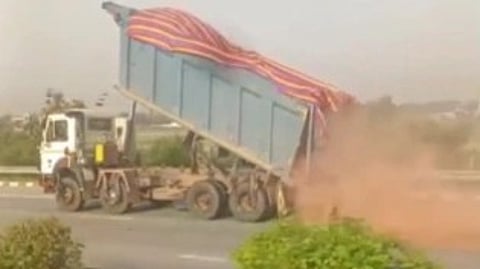 A truck is tilted on a highway, dumping a load of red dirt. Dust clouds arise as vegetation lines the road. The scene conveys motion and activity.