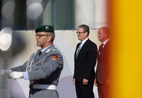 Image of Prime Minister Keir Starmer meets German Chancellor Olaf Scholz in Berlin, Germany. The extreme right side of the image has a orange yellow shade . there are three people in the image with starmer standing sideways in the center.