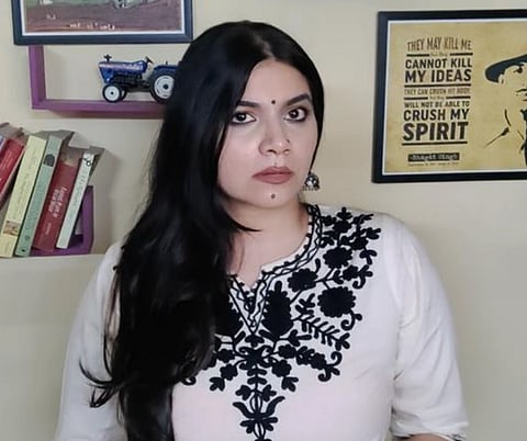 Image of Ruchika Sharma standing behind a wall with book shelf and a frame of Bhagat Singh.