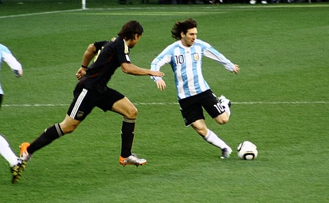 Image of Argentina’s Lionel Messi playing on the ground. the image is older as Messi has longer hair and is kicking the ball with his left foot.