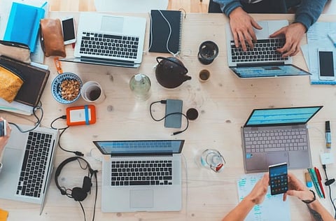Image of a group of people sitting on a table with their laptops, phones and headphones placed on it. the image is taken from above.