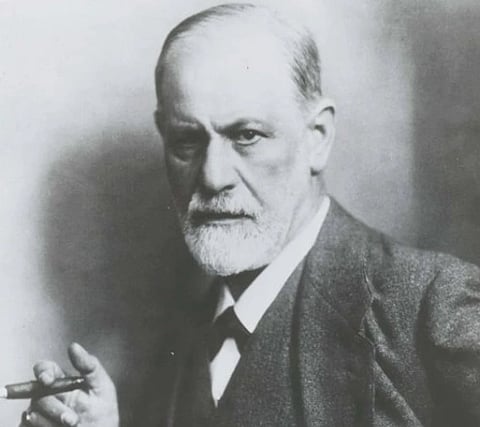 An older man with a serious expression holds a cigar in his right hand. He's dressed in a suit and tie, set against a softly blurred background.