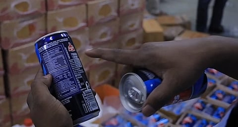 image is an screenshot from a video where a man is at a food godown/ warehouse and he is holding a soft drink cans in his hands. he is pointing his finger towards the ingredient panel of the can