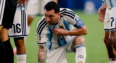 Image of Lionel Messi sitting halfway on the ground. his one hand is placed on the grass. Tow three people are standing with him.