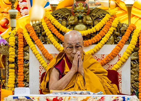 Image of Dalai Lala with his hands folded and seated in a chair decorated with flowers.