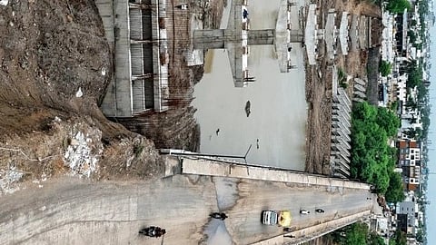 Aerial view of a partially constructed bridge over a river. Cars and motorcycles navigate the completed section. Urban area with buildings and greenery surrounds.