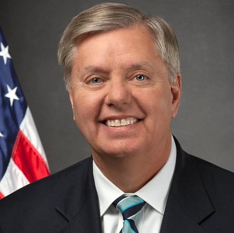 Image of late US senate Lindsey Graham smiling and behind him is the flag of USA.