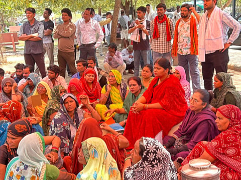 Image of a group of people in a rural India sitting together. they are all wearing sarees and suits. Behind them a group of men are standing.