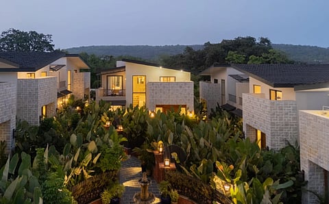 Modern villas with stone facades and glowing windows are nestled among lush greenery at dusk. A peaceful, inviting atmosphere is created by the soft lighting and surrounding trees.