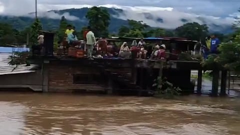 In the image Assam flood is shown with people standing on the top of the house